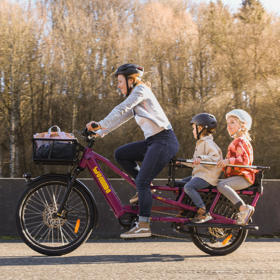 Eine Frau transportiert ihre Kinder mit Yuba Spicy Curry in Farbe very berry anm See entlang.